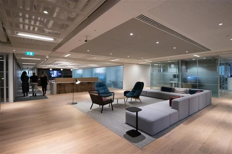 Ashurst's contemporary office interior with open lounge area, modern furniture, wooden floors, and glass-walled meeting rooms, with three people walking through the corridor.